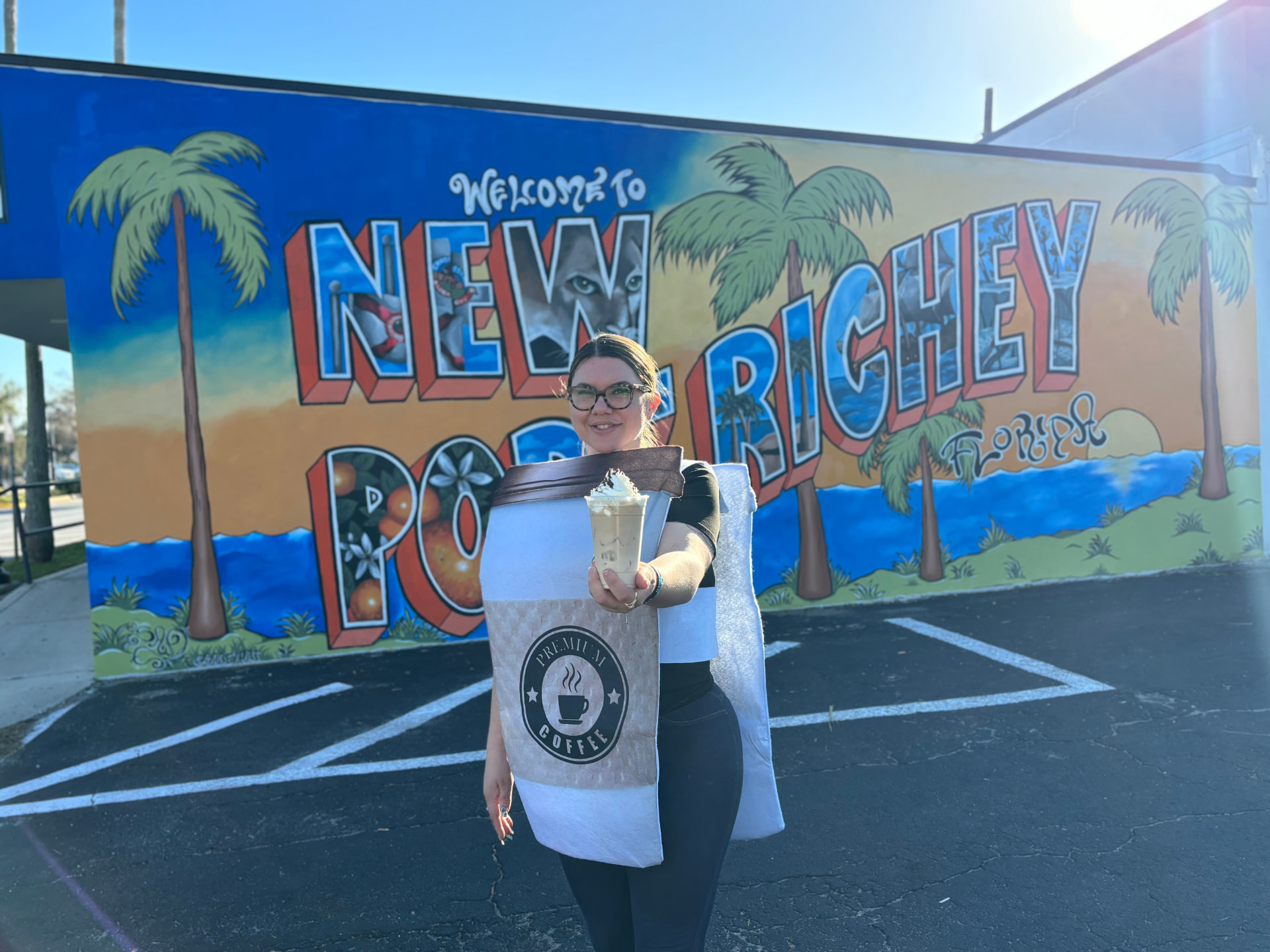 Sarah, owner of The Skinny Bean Cafe, holding an iced latte in front of the Welcome to New Port Richey mural